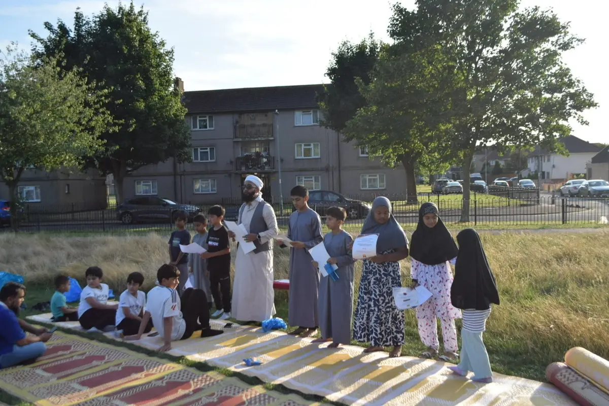 Children standing and sitting on mats outdoors, holding certificates, with a teacher beside them